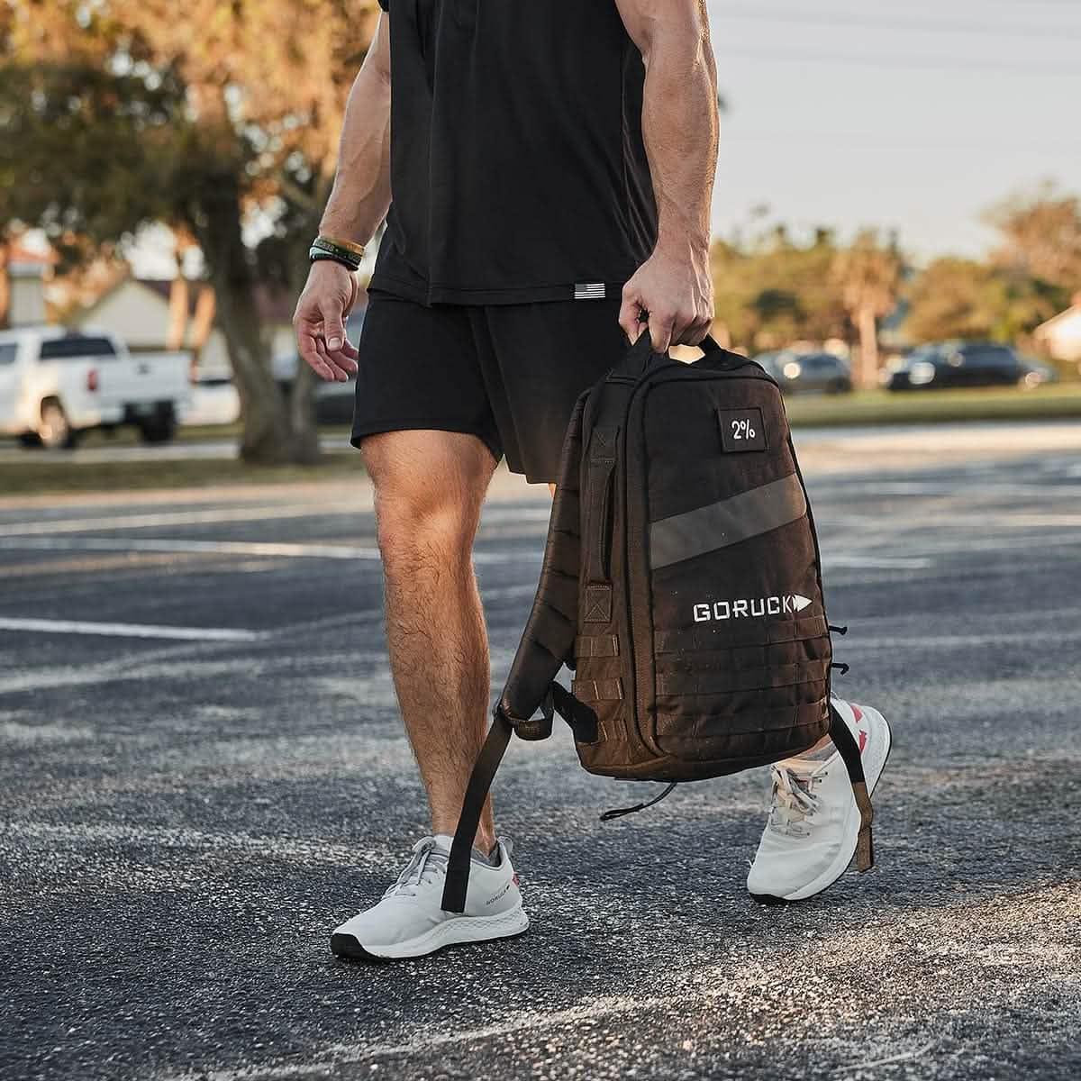 Person in black athletic wear holding black GORUCK backpack outdoors on pavement