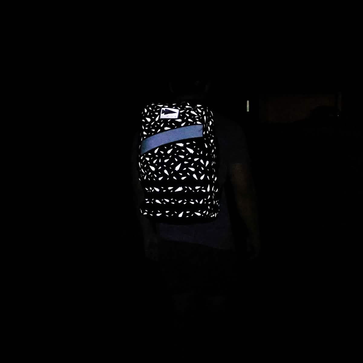 A person in darkness wears the Rucker 4.0 - Reflective Spearhead, a black rucksack featuring white reflective patterns and a blue diagonal stripe.