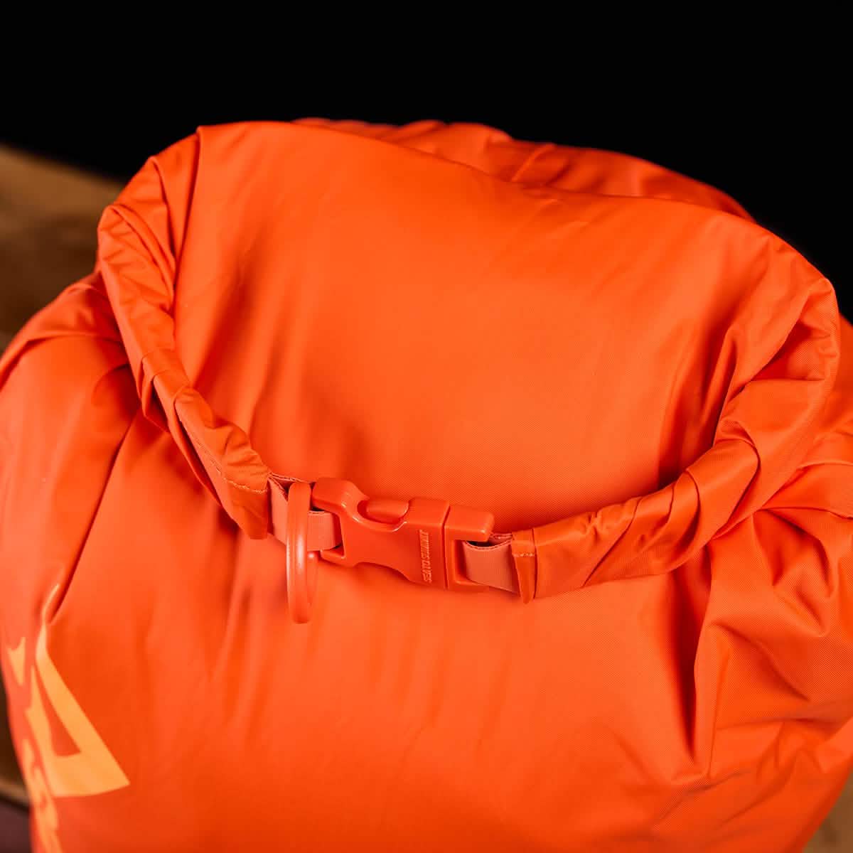 A close-up of the Sea to Summit Dry Sack crafted from lightweight orange nylon showcases a roll-top waterproof closure secured with a plastic clip. The material appears smooth and durable, ideal for keeping contents dry during outdoor activities. The dark background accentuates the bag's vibrant color.
