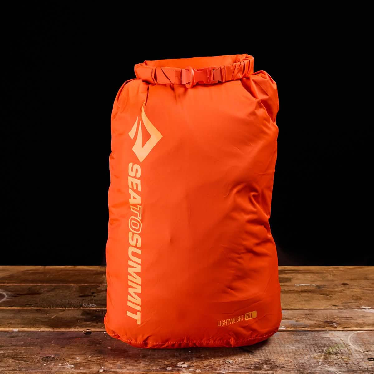 A vivid orange Sea to Summit Dry Sack, made from lightweight nylon, is positioned on a wooden surface with a black background. The sack boasts a waterproof roll-top closure and displays the brand's name in white text along the side.