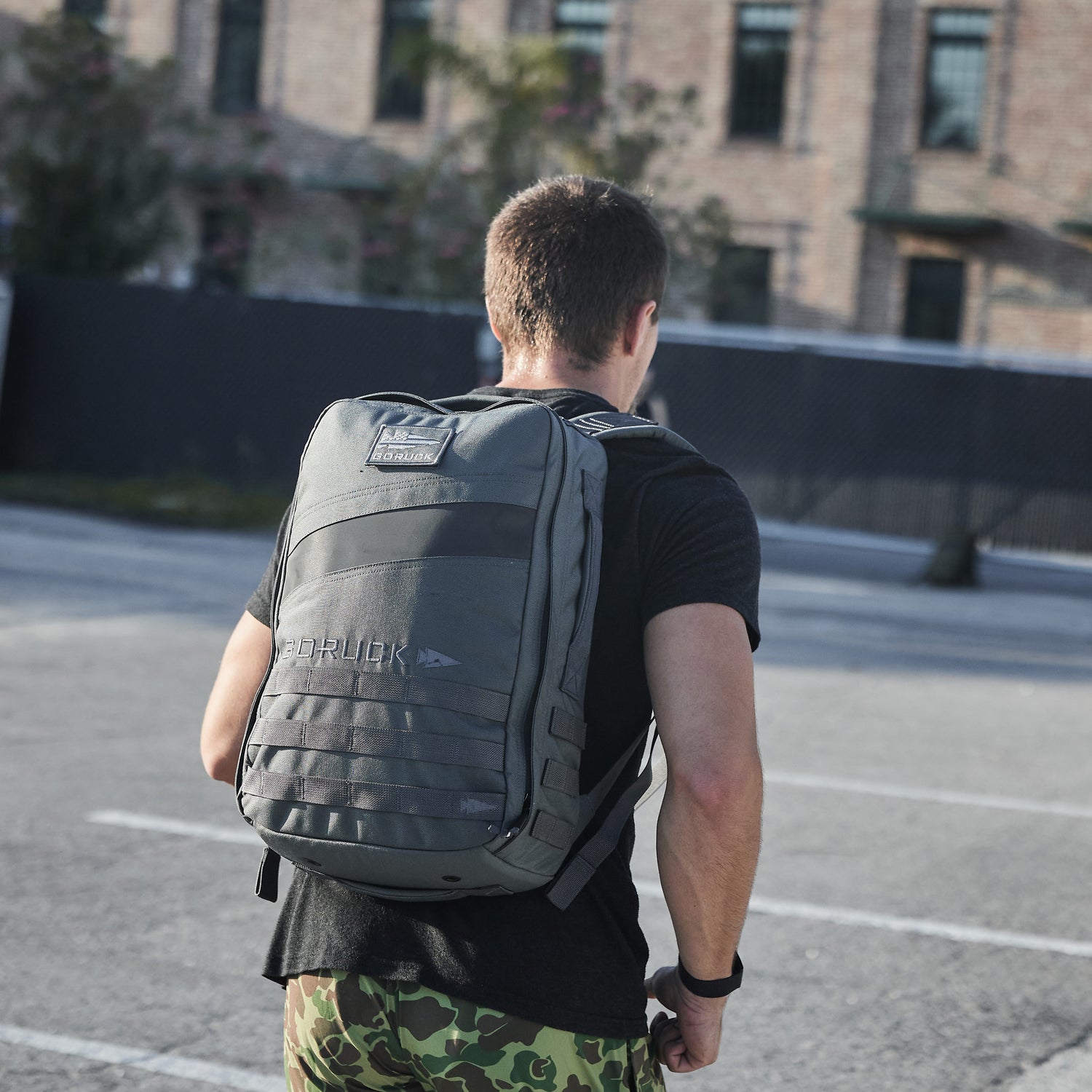 A man wearing the Rucker 4.0, a large gray rucksack, stands in an empty parking lot, facing away from the camera—possibly using the space as a training tool for rucking.