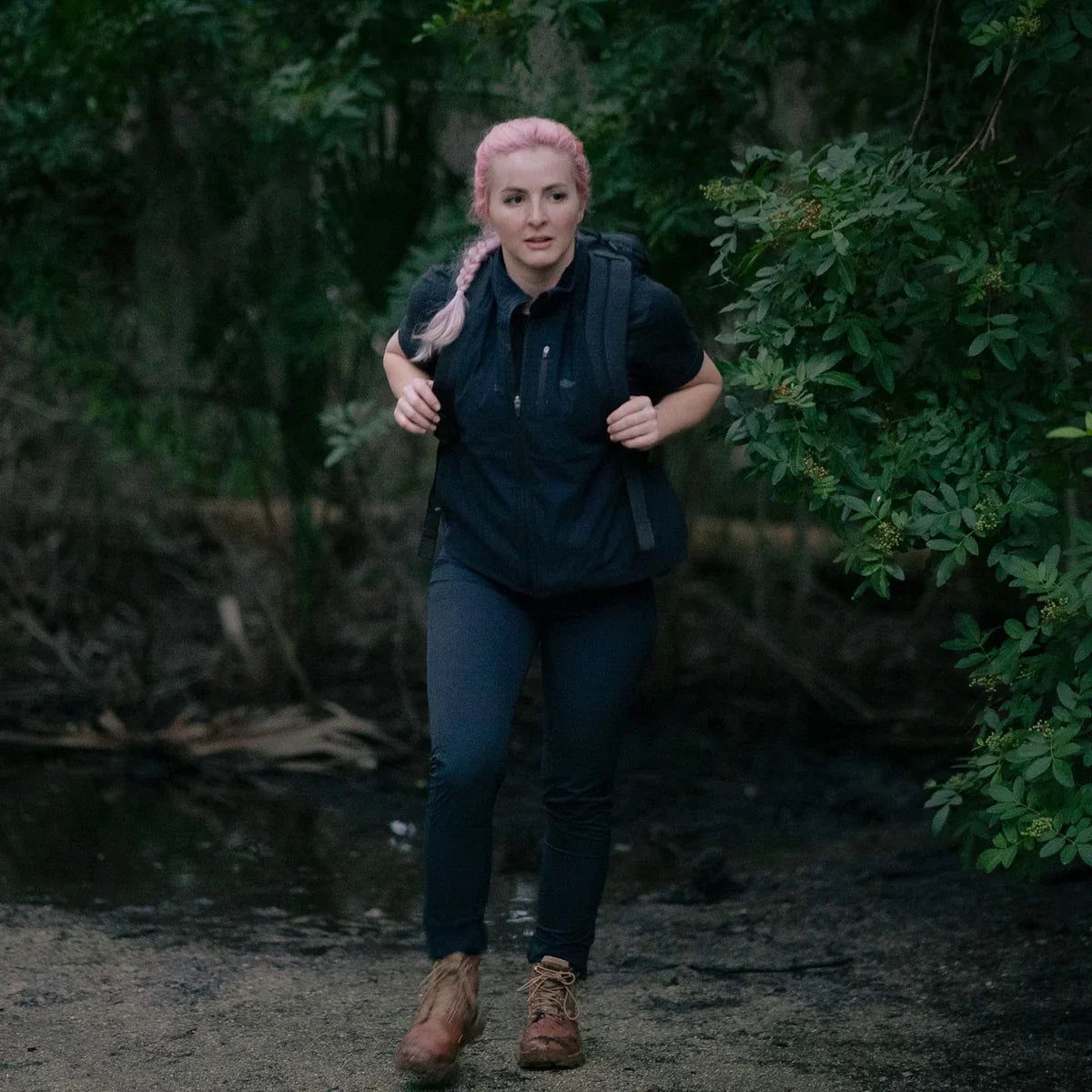 Woman with pink braided hair hiking through forest wearing black activewear and a GORUCK backpack