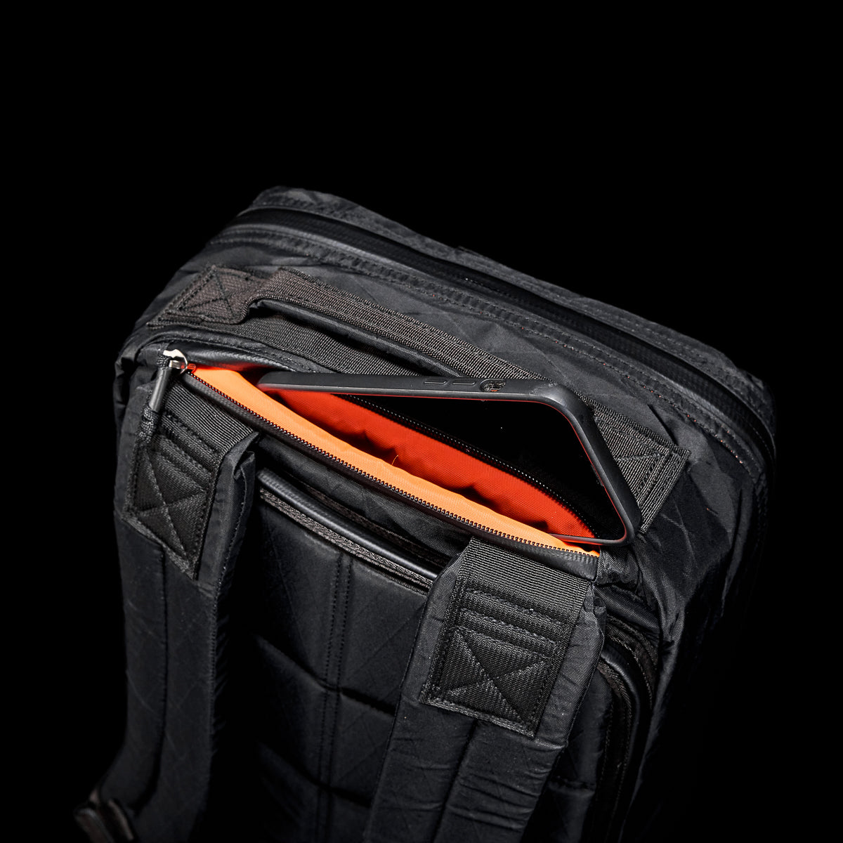 The GR1 USA - X-PAC rucksack, featuring a black exterior and orange interior, is partially open to reveal a smartphone against a black backdrop.
