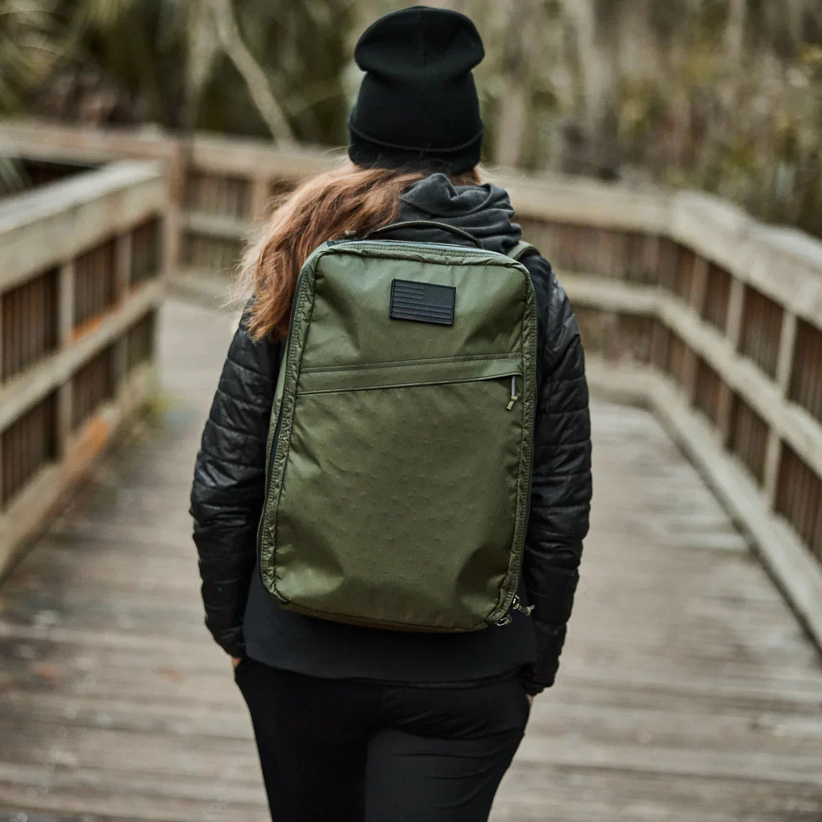 Person walking on wooden bridge wearing black jacket and beanie with green GORUCK rucksack backpack