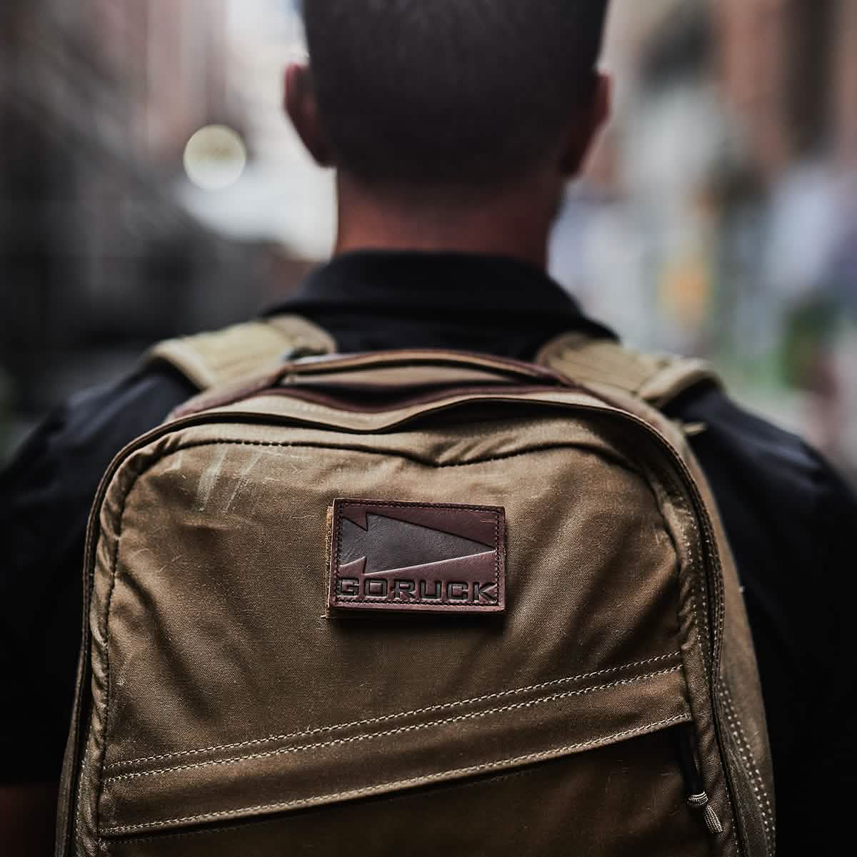 A person faces away from the camera, displaying the GORUCK GR1 USA - Heritage backpack made from waxed duck canvas. The visible GORUCK logo patch adds to its appeal. A blurred city street behind them emphasizes the durable and stylish characteristics of this rucksack.