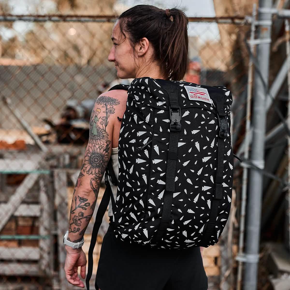 Woman with a tattooed arm wearing a black and white patterned M23 - Ballistic Nylon Cordura rucksack, standing near a wire fence—the ideal durable everyday carry for urban adventures.