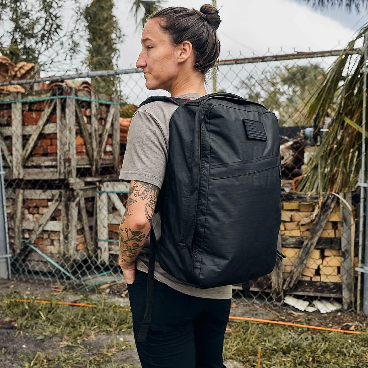 Person with tattoos wearing a black GORUCK backpack outdoors near a chain-link fence and stacked wood