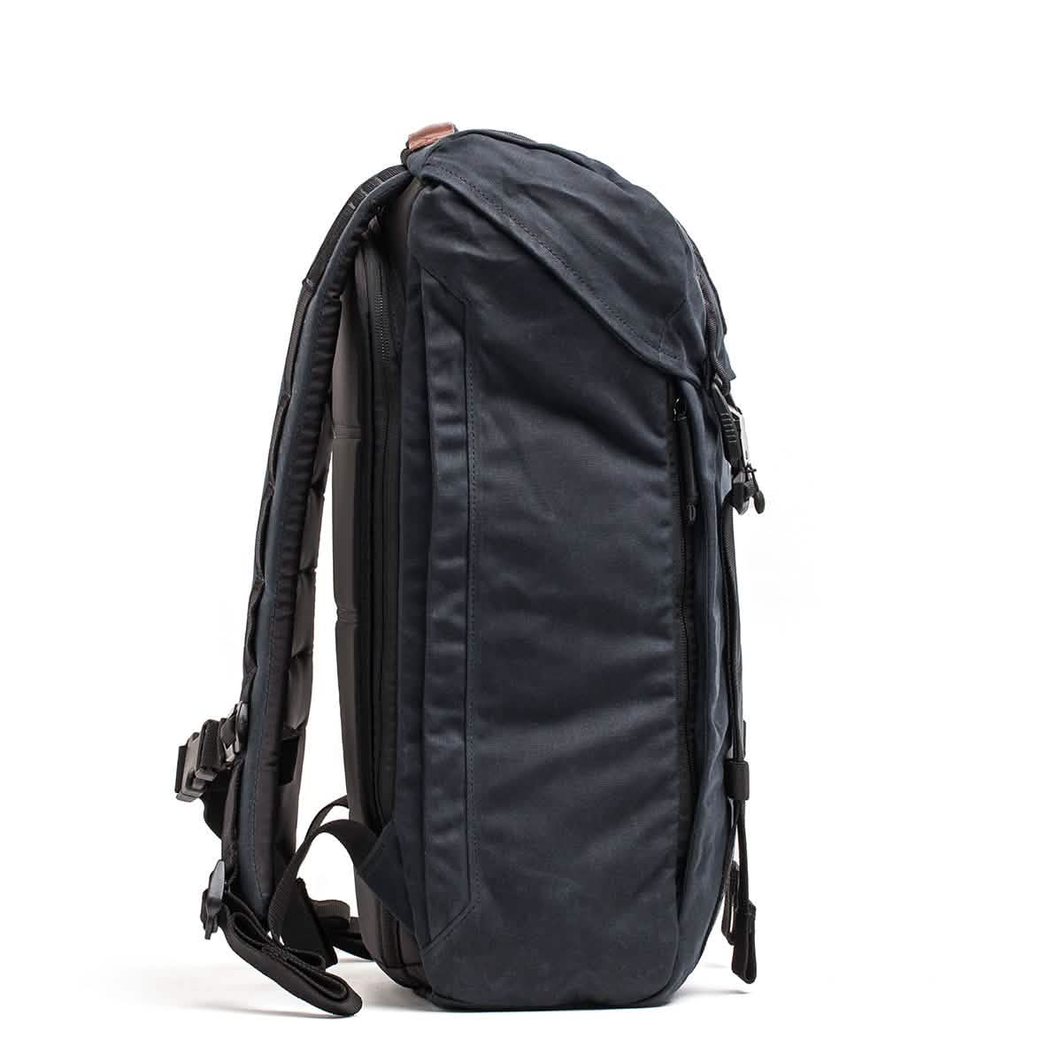 The GORUCK M23 - Waxed Canvas is a stylish black rucksack featuring adjustable straps and several zippered compartments, displayed from the side. Made from durable materials, its design incorporates a curved, structured back for added comfort and support.