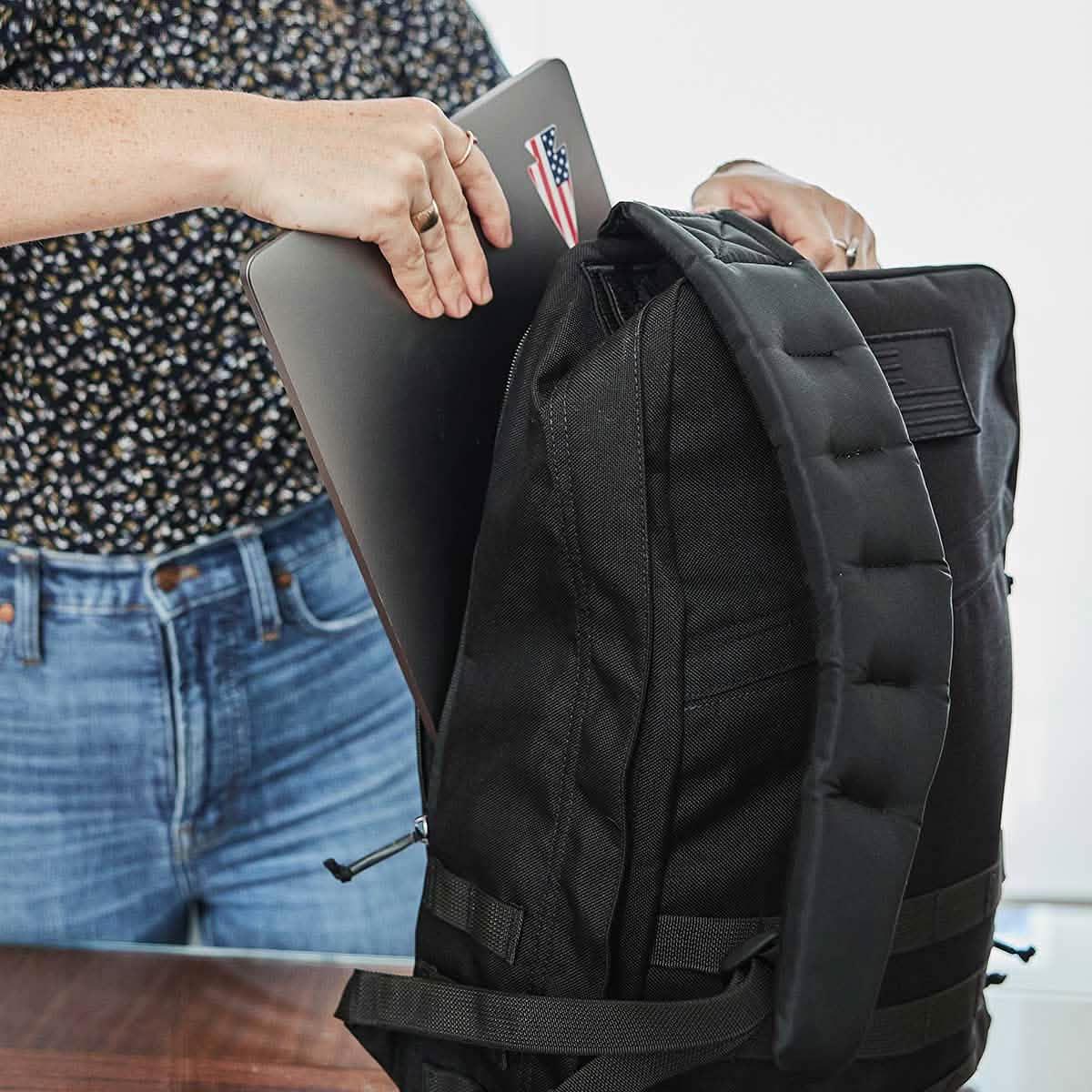 Person placing laptop with American flag sticker into black GORUCK durable backpack