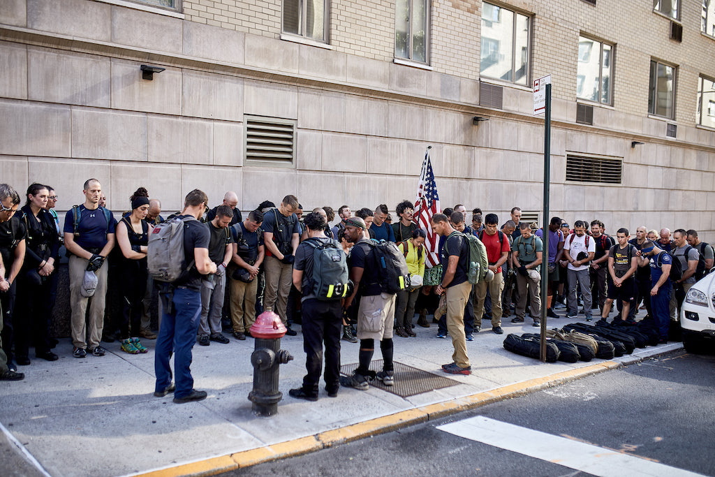 A group of people with backpacks stand and bow their heads on a city sidewalk, holding an American flag.
