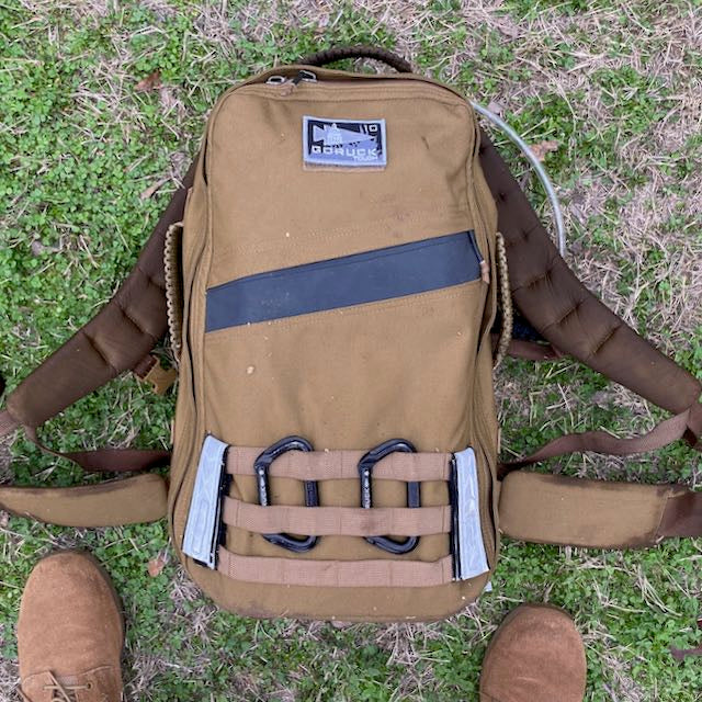 A tan GORUCK backpack with two black MOLLE rucksack carabiners attached to external webbing, resting on grass near a pair of brown boots.