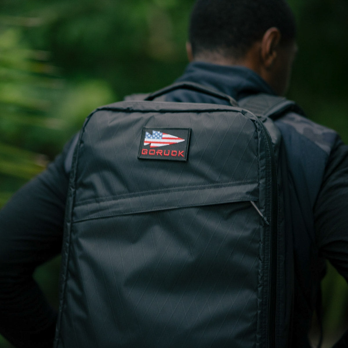 A person wearing a GR1 USA - X-PAC rugged black backpack with an American flag patch and AquaGuard zippers for waterproofing stands out against lush greenery.