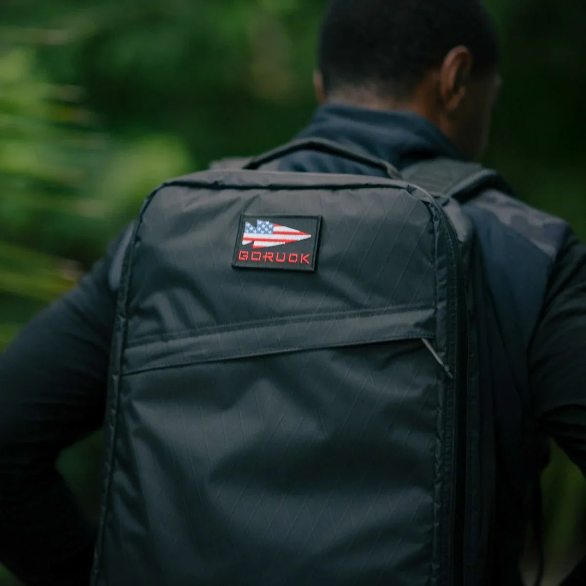 Close-up of a person wearing a black GORUCK backpack with an American flag patch in an outdoor green setting