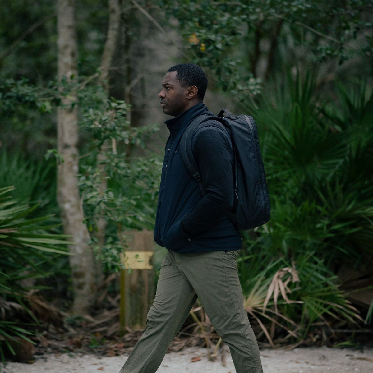 A man strolls through the forest, his dark jacket complemented by a GR1 USA - X-PAC backpack featuring durable fabric and AquaGuard zippers. With hands in pockets, he blends seamlessly into the lush greenery.