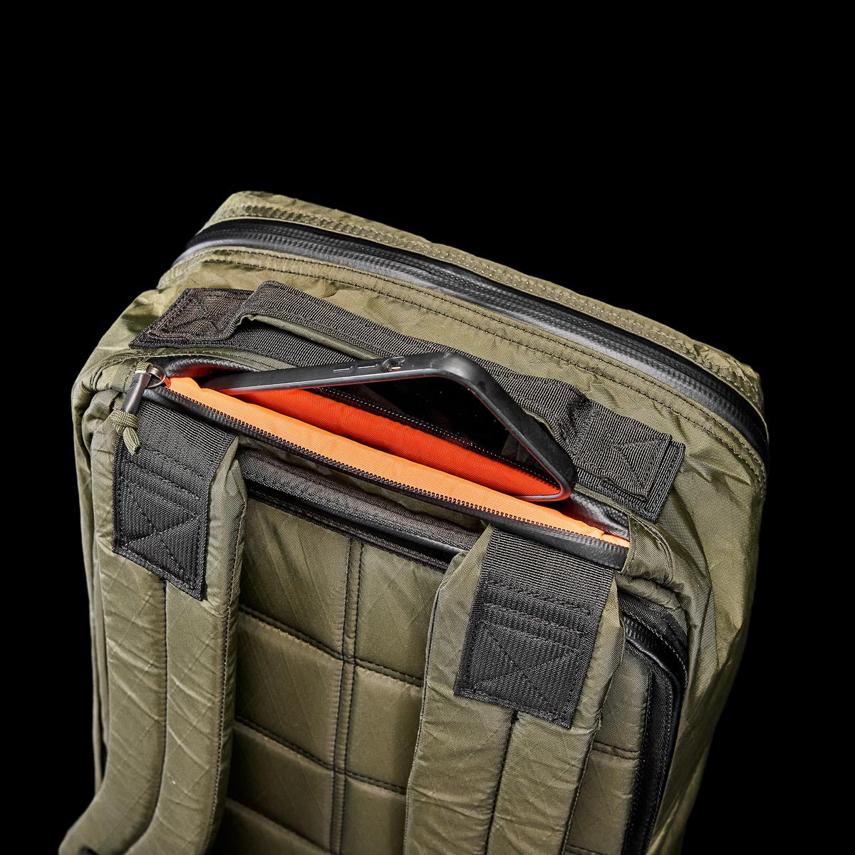 An olive green GR1 USA - X-PAC rucksack, showcasing a tablet in the top pocket, designed from durable X-PAC material against a black backdrop.