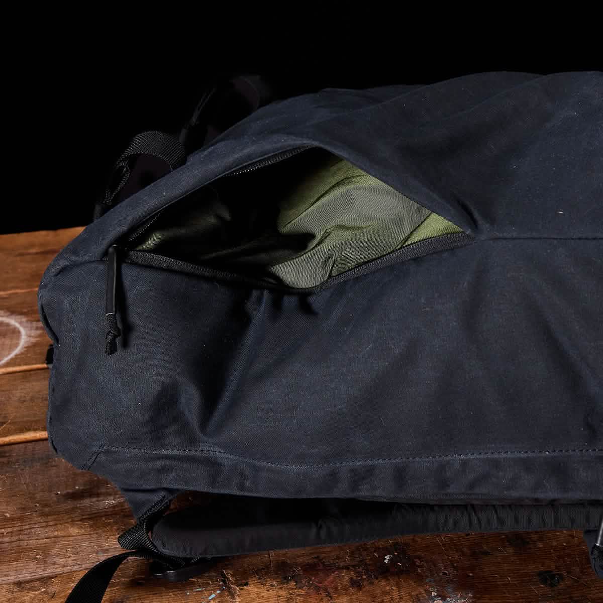 A black backpack, similar to the M23 - Waxed Canvas from GORUCK known for its standard durability, features a partially open zippered pocket with green fabric lining peeking out. It is displayed on a rustic wooden surface against a dark backdrop.