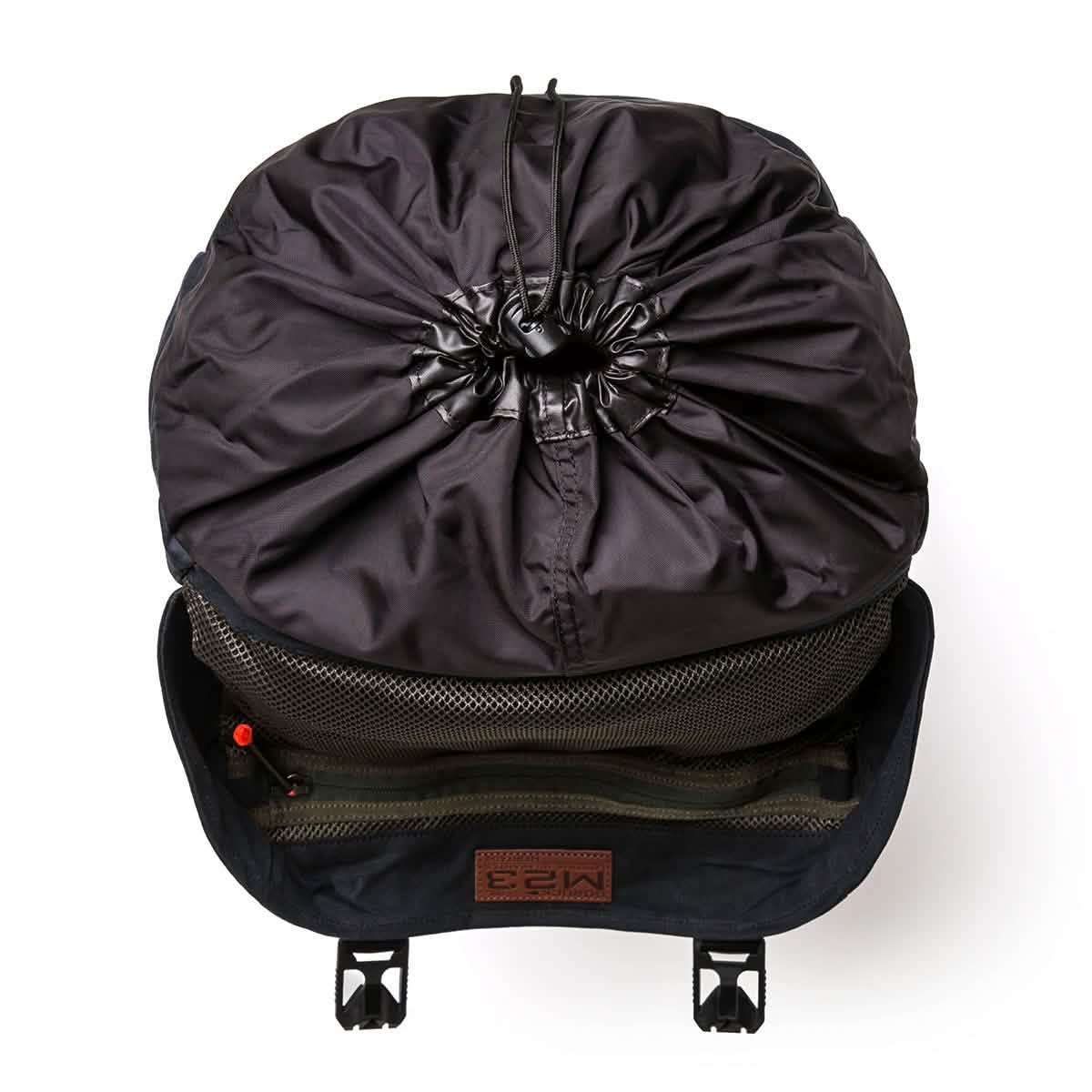 A black modern rucksack from GORUCK's M23 - Waxed Canvas collection is viewed from above, highlighting its drawstring closure at the top. The partially open interior reveals organizational pockets and a patterned fabric lining, with two small buckles visible at the bottom edge.
