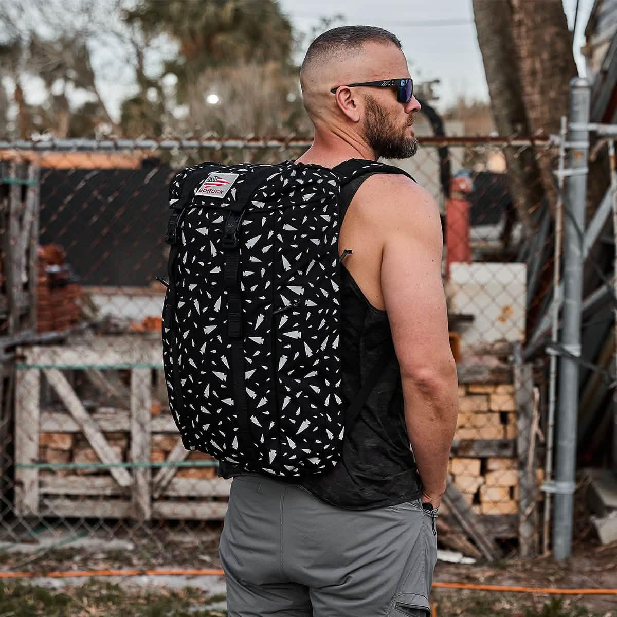 A man in sunglasses and a tank top wears the M23 - Ballistic Nylon Cordura, a black and white patterned backpack, outdoors near a fence.