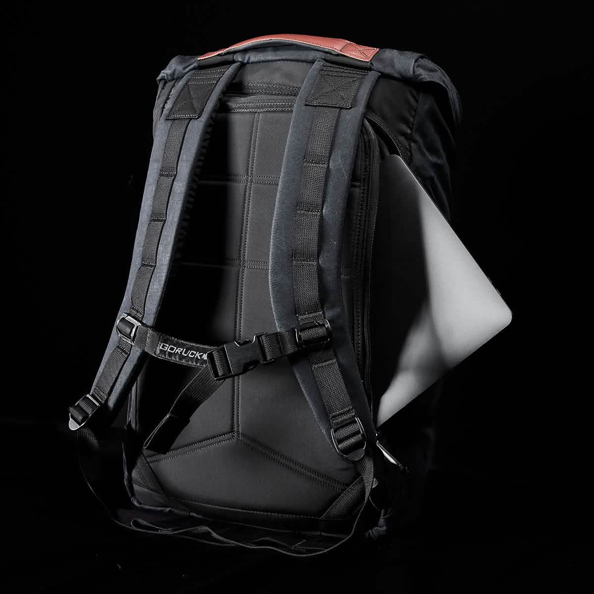 Against a dark background, the GORUCK M23 - Waxed Canvas backpack is prominently displayed, with a white laptop partially visible from the side. The image emphasizes its back and padded shoulder strap design.
