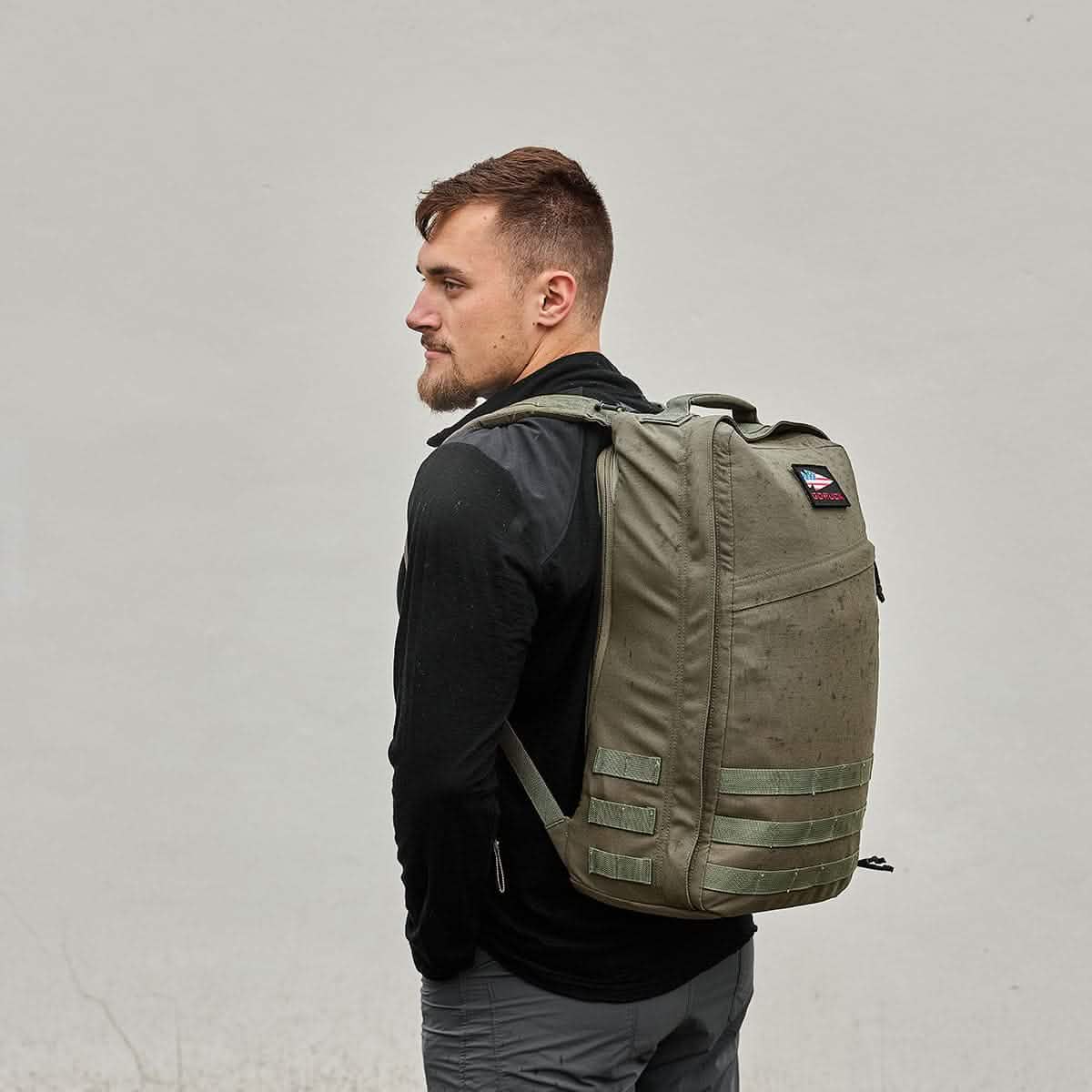 A person with short hair stands sideways against a light gray background, wearing a black jacket and carrying a large olive green GR1 USA - Cordura backpack by GORUCK, perfect for the GORUCK Challenge.