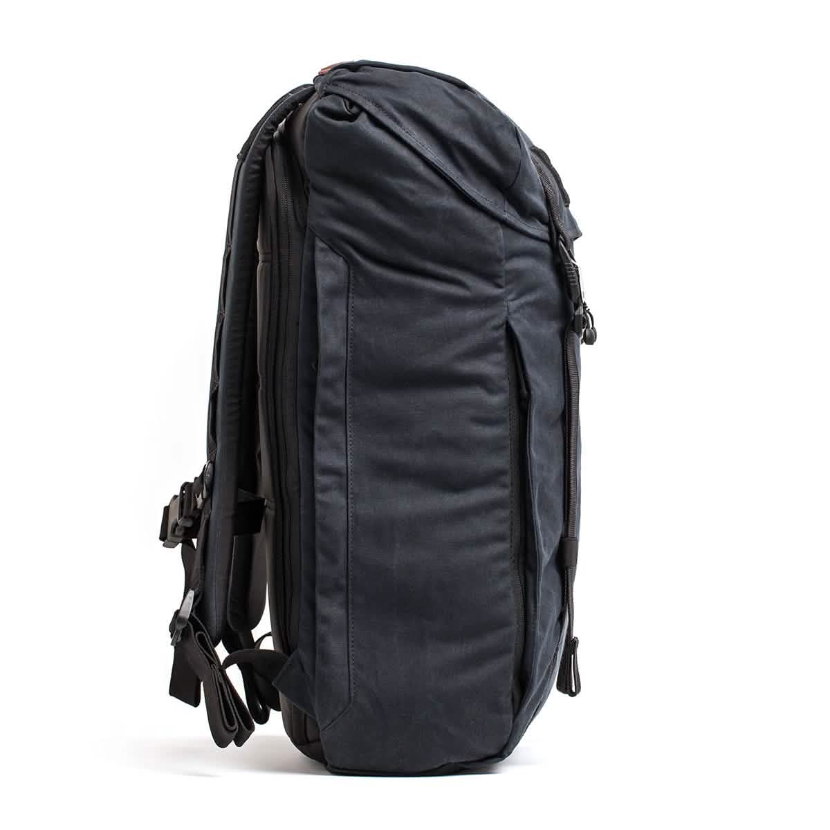 The GORUCK M23 - Waxed Canvas rucksack, depicted in a sideways view, comes in dark gray and features multiple compartments along with padded shoulder straps. Its streamlined design is accentuated by buckles, providing a military-inspired style that's perfect for travel or daily use.