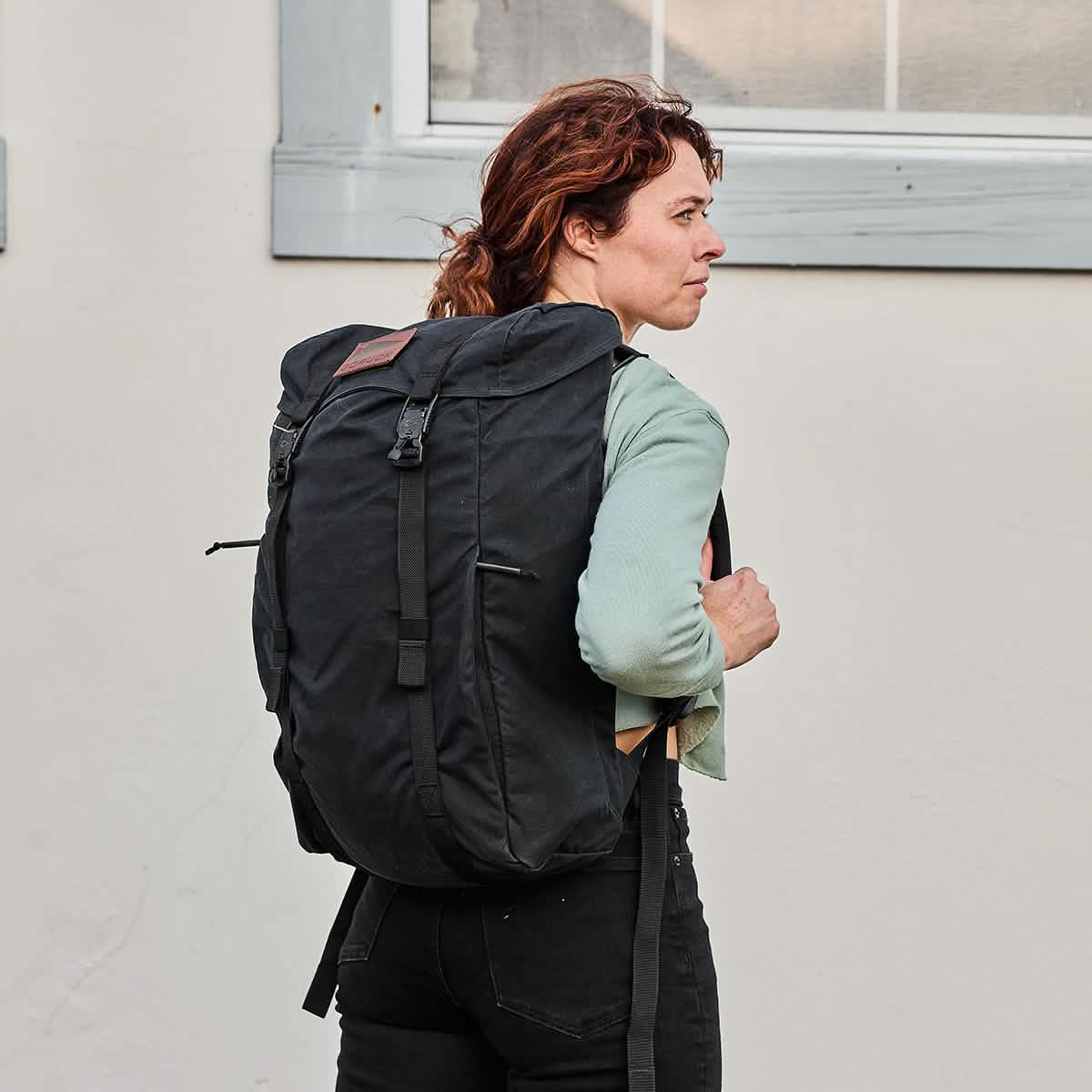 A person with curly hair, dressed in a green shirt and black pants, is facing left while carrying a GORUCK M23 - Waxed Canvas rucksack in front of a light-colored wall with a window.