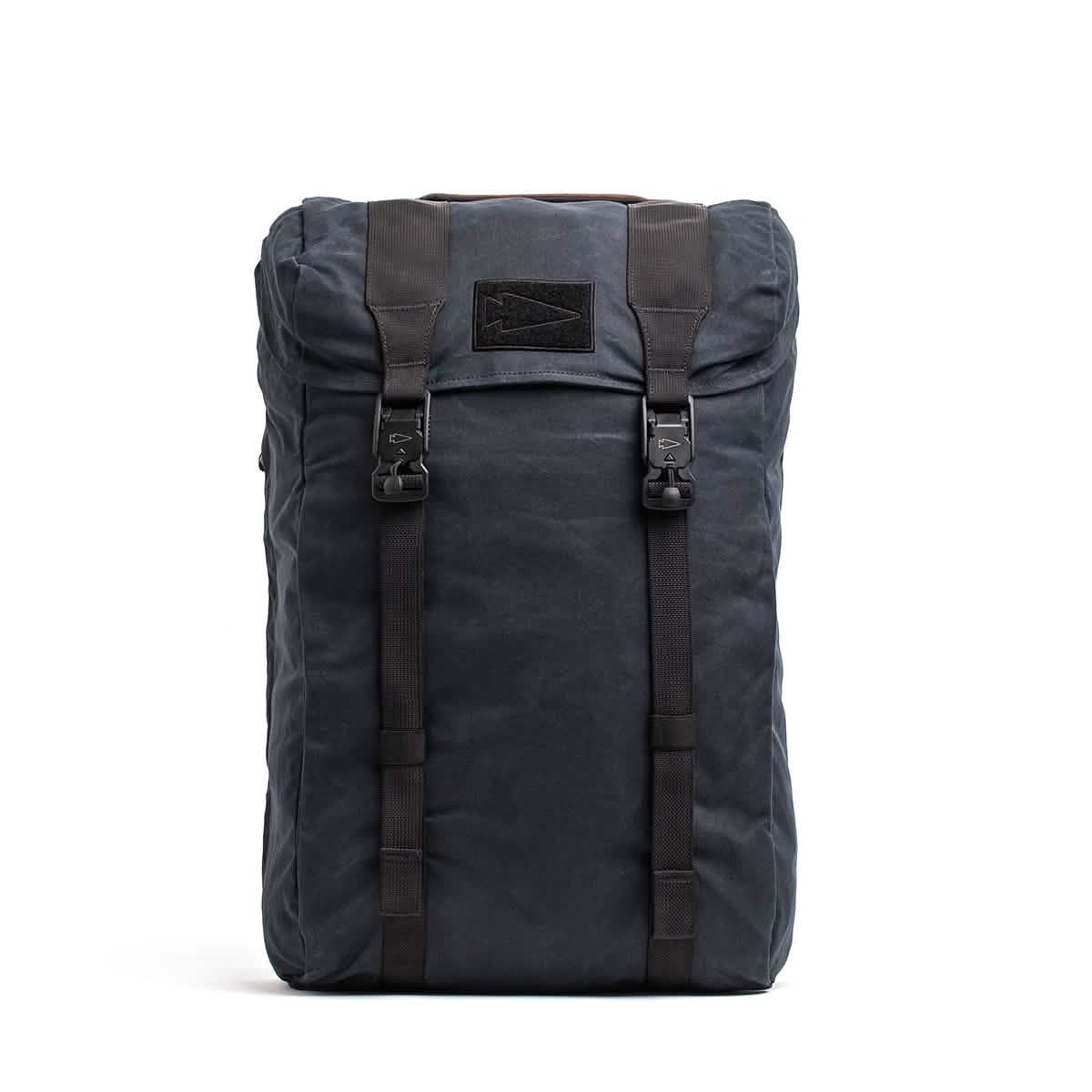 The M23 - Waxed Canvas by GORUCK is a military-inspired rucksack in dark gray, featuring two black straps and buckles on the front, a top flap closure, and a rectangular patch in the center. Its simple yet rugged design makes it perfect for outdoor use.