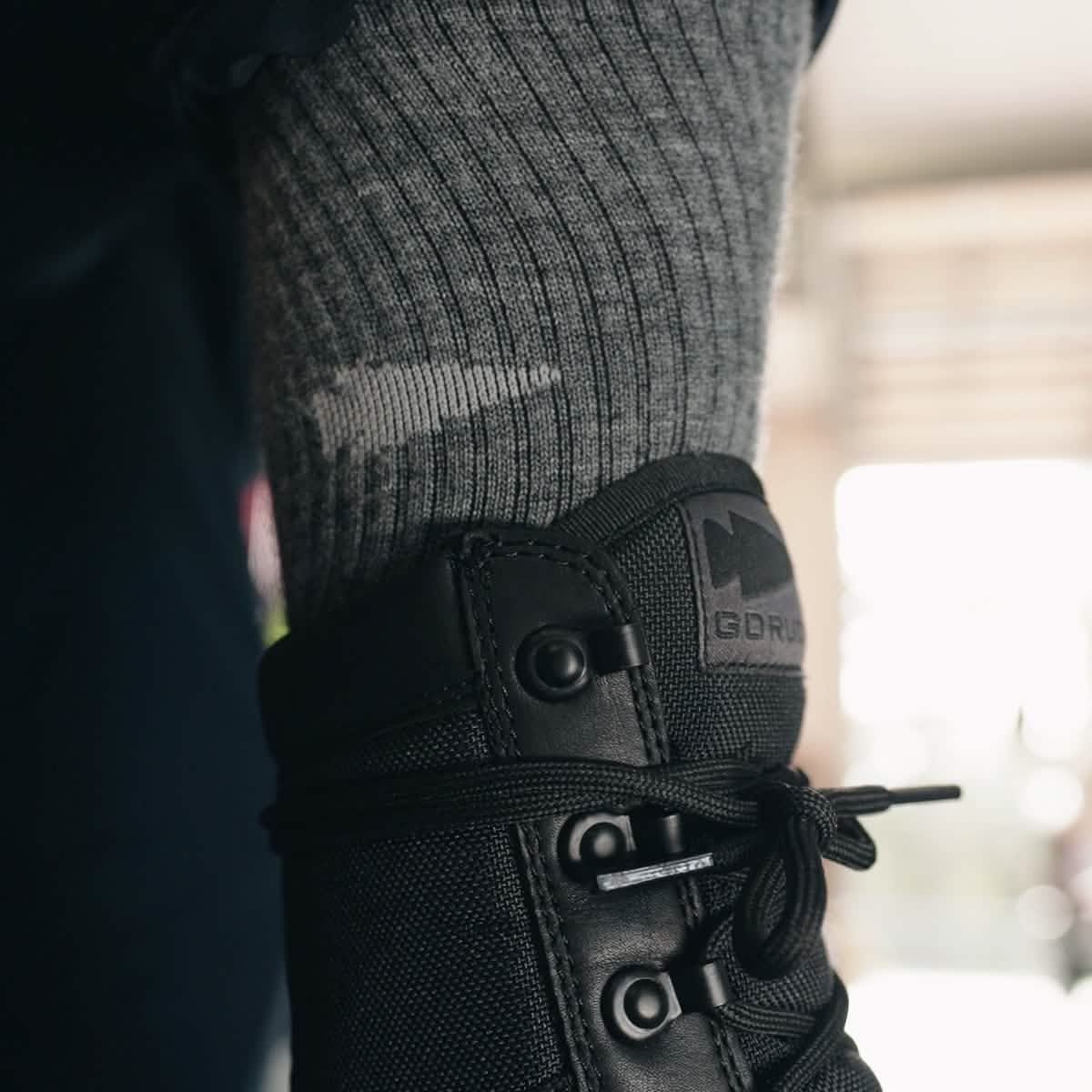 Close-up of a foot wearing a grey, ribbed sock with a small logo inside a black MACV-2 Safety Boot - High Top by GORUCK meeting Special Forces standards.