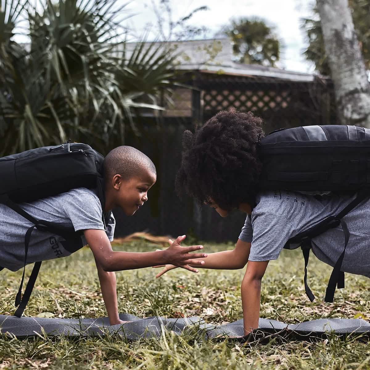 In a grassy area surrounded by palm trees, two children participating in the GORUCK Elementary School Ruck Training Program are in a garden plank position, high-fiving each other with their free hands as they master functional movements while wearing backpacks.