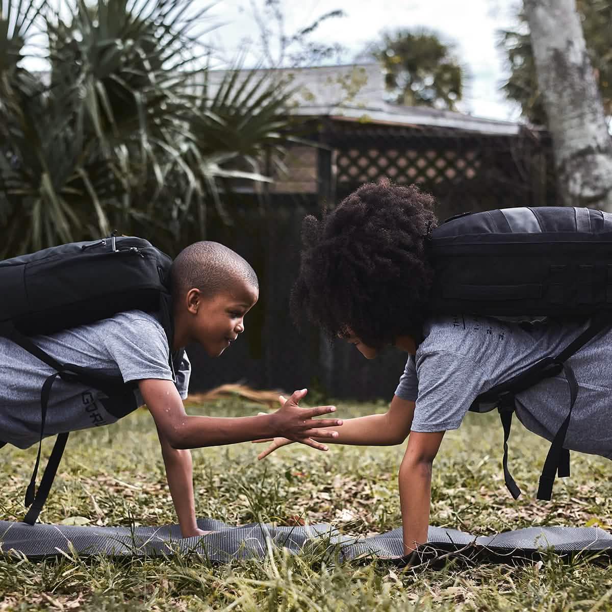 Two kids outdoors doing plank exercises wearing GORUCK backpacks and gray shirts, high-fiving each other on mats