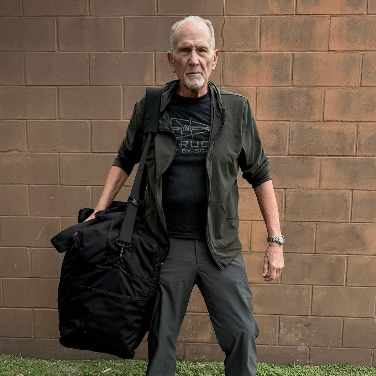 Older man wearing GORUCK shirt and jacket holding large black ruck bag outdoors