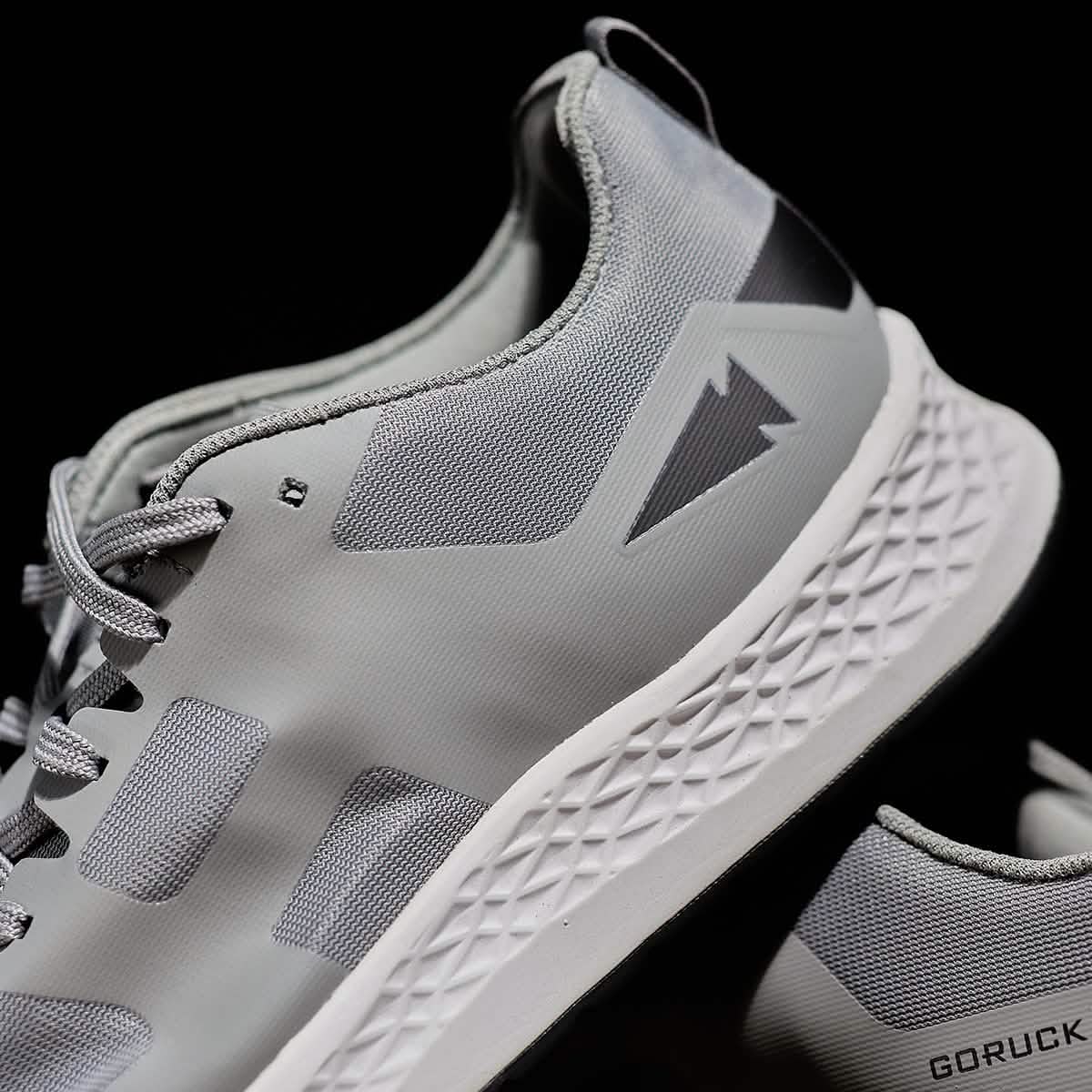 A close-up of the Men's Rough Runner - Speed Grey + White athletic shoe, featuring a gray design with a white textured sole, perfect for the Rough Runner. This GORUCK shoe incorporates mesh fabric and subtle details, along with Gradient Density technology, and displays a triangular logo. The brand name is prominently visible on the heel against a dark background.
