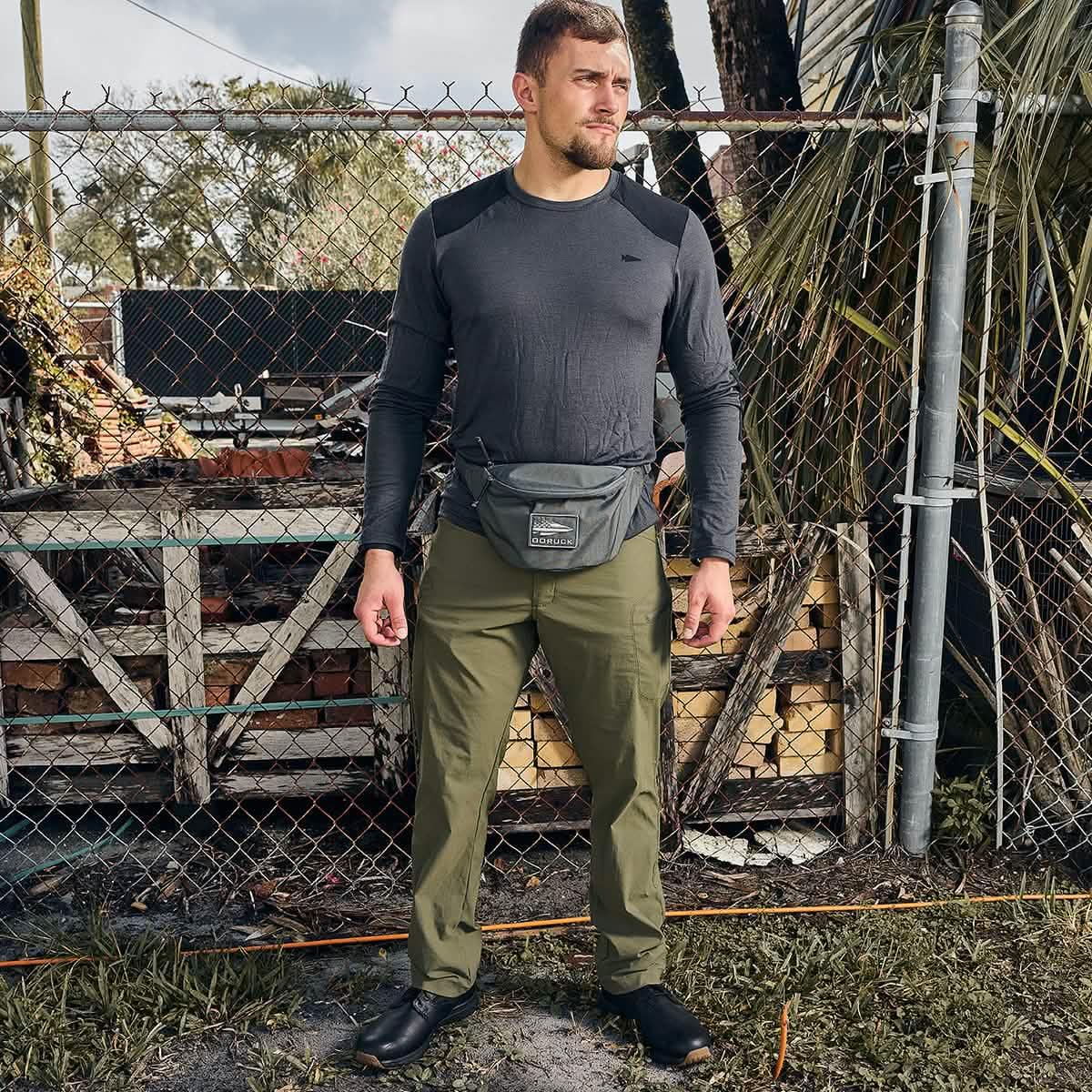 Man wearing GORUCK gear with a gray chest rig and olive pants outdoors by a fence