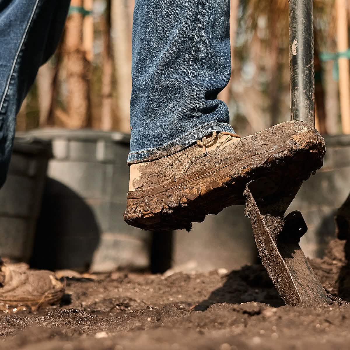 A close-up of a person wearing the GORUCK MACV-2 Safety Boot - Mid Top and jeans, stepping on a shovel in a garden or outdoor setting. Large plant pots can be seen in the background.
