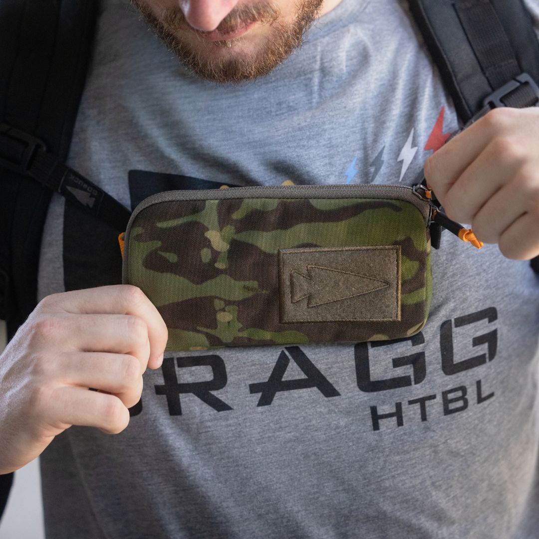 A person holds the Sternum / Belt Pouch - CORDURA in camo on their chest while wearing a gray shirt with “FRAGG HTBL” text, keeping their essentials hands-free—ideal for rucking or outdoor adventures.