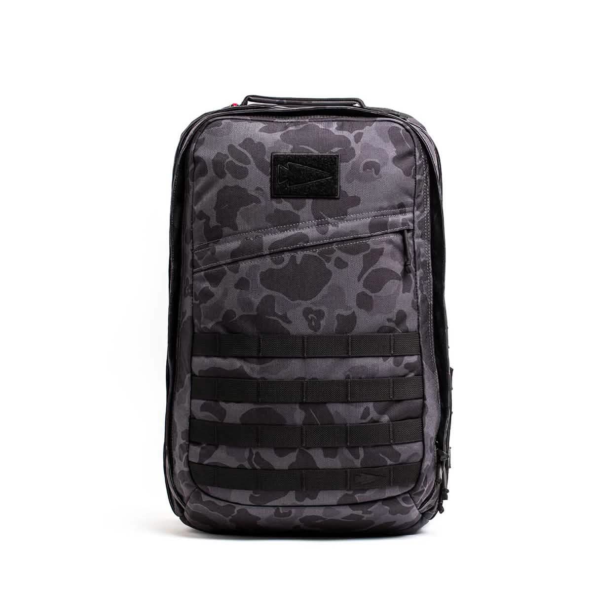 A GORUCK GR2 backpack in gray camouflage with black straps and multiple compartments stands upright against a plain white background. The design, inspired by Special Forces rucks, features horizontal webbing on the front.