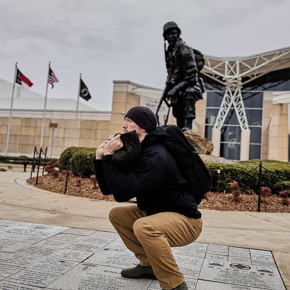 Man wearing GORUCK gear performing sandbag squat outdoors near military statue and flags.