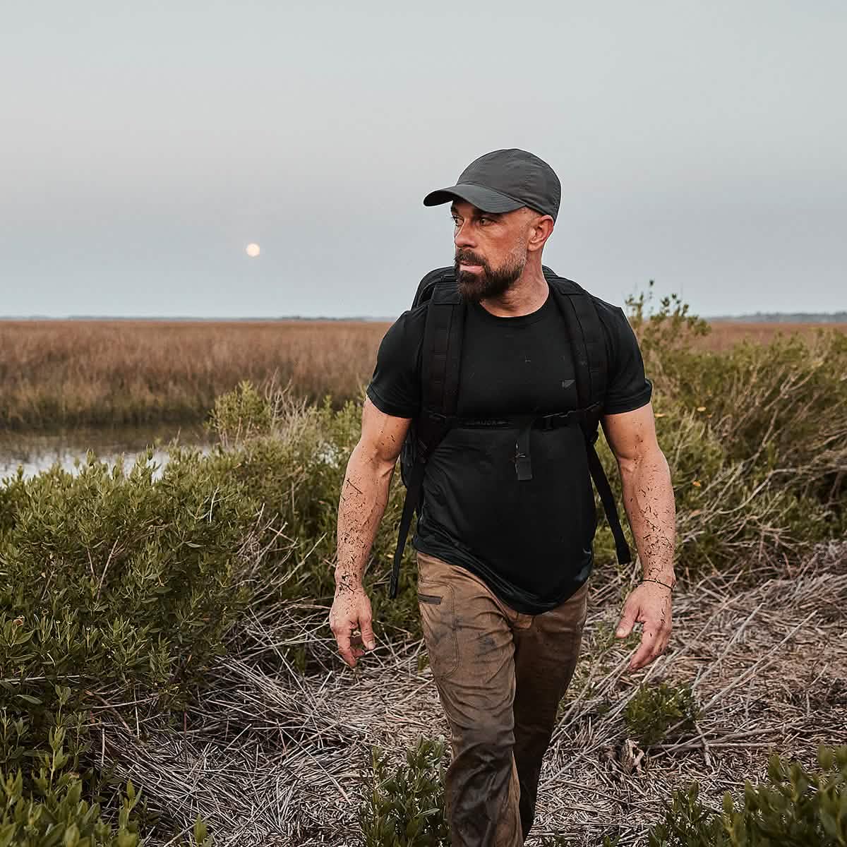 A man in outdoor clothing and a cap, carrying the Rucker - Long Range rucksack, walks through marshy grassland at dusk with a serious expression.