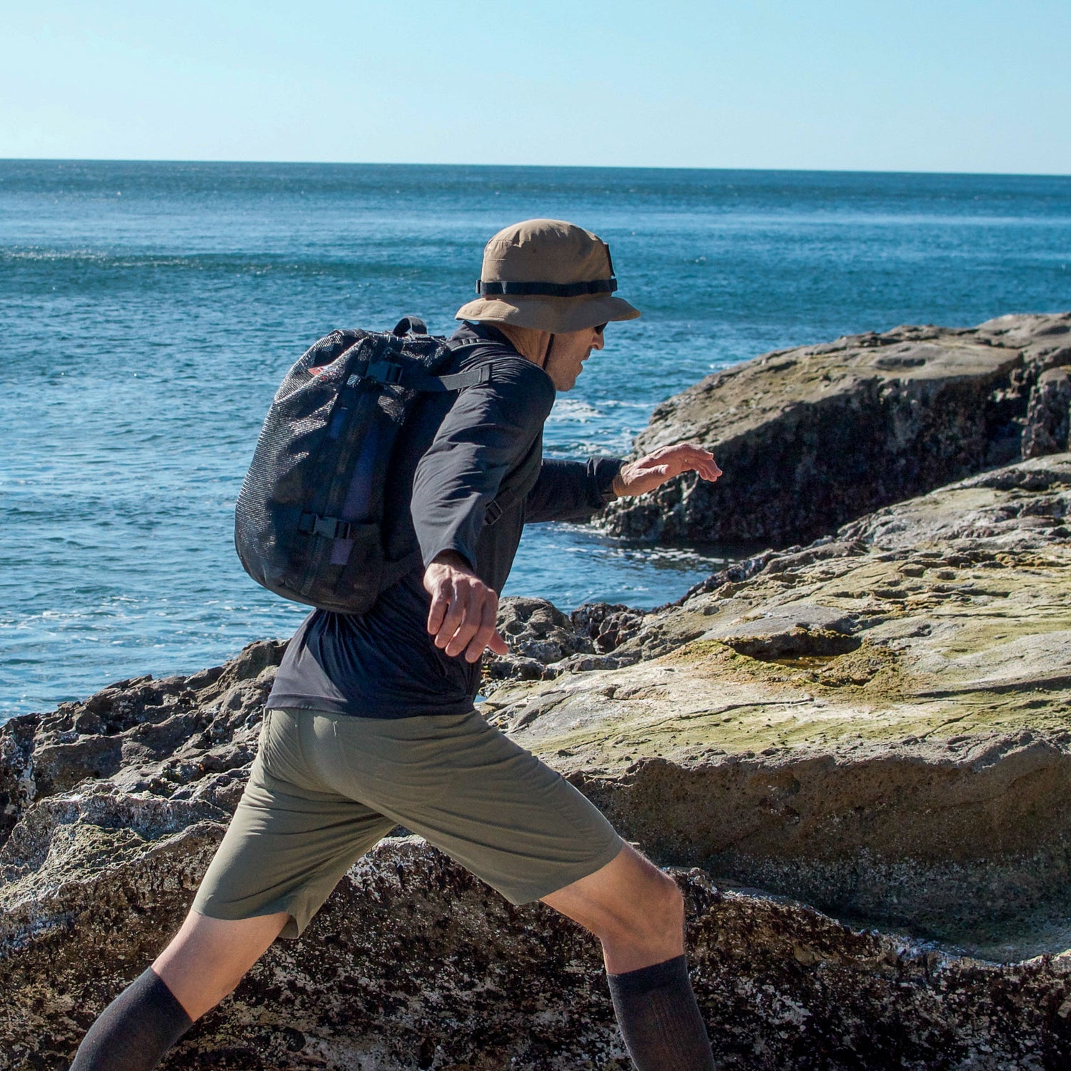Carrying the Mesh Ruck, someone treks across rocky coastal terrain beneath a clear blue sky.