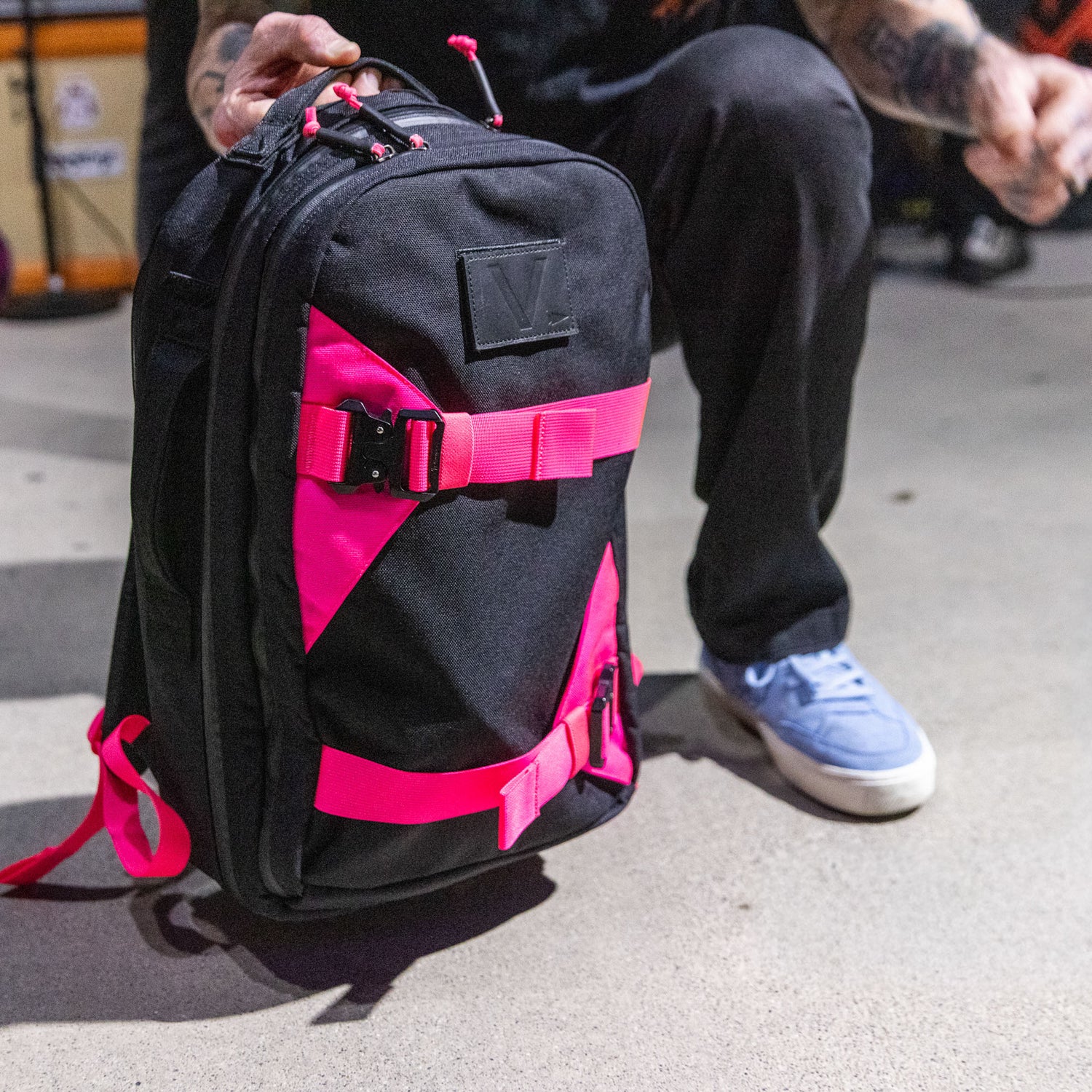 A person kneels on a concrete floor, holding the Skate Ruck with bright pink straps and a durable laptop compartment.