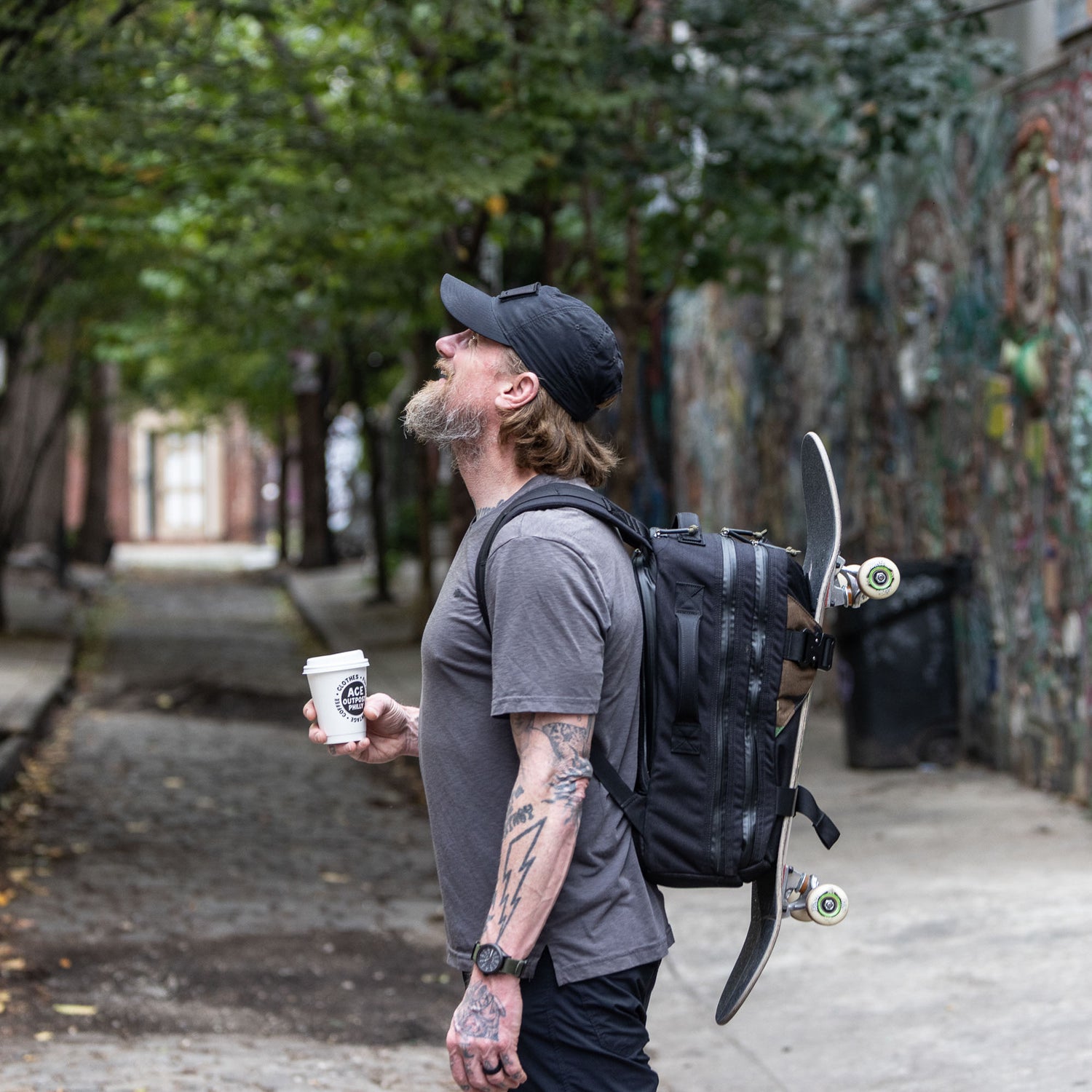 A tattooed man carrying a Skate Ruck bag with a bombproof laptop compartment and coffee stands in a graffiti-covered alley, looking up.