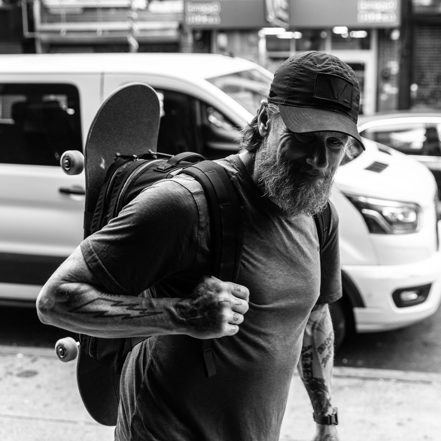 A tattooed, bearded man carries his Skate Ruck, which features a bombproof laptop compartment and holds a skateboard, as he walks down a city street with a van in the background.