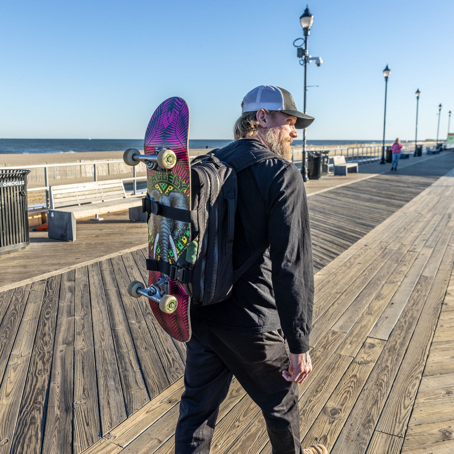 A man walks along a sunny beach boardwalk, carrying his skateboard on his Skate Ruck—a bag with a bombproof laptop compartment—enjoying the day with his essential EDC.