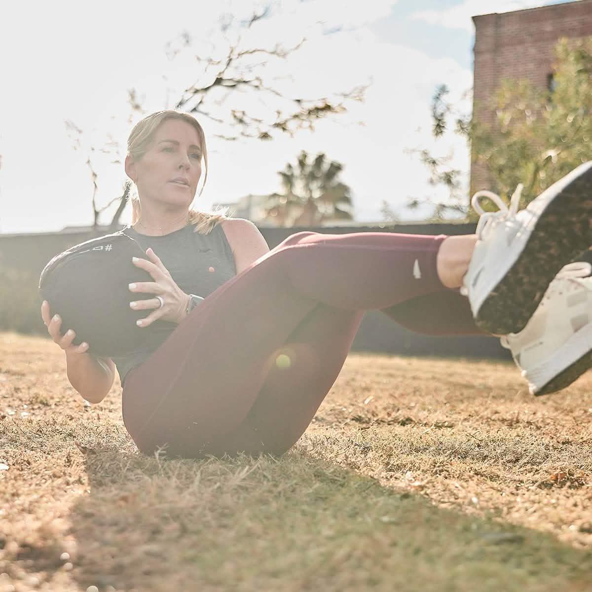 Woman exercising outdoors with a weighted sandbag wearing athletic gear and white sneakers on dried grass