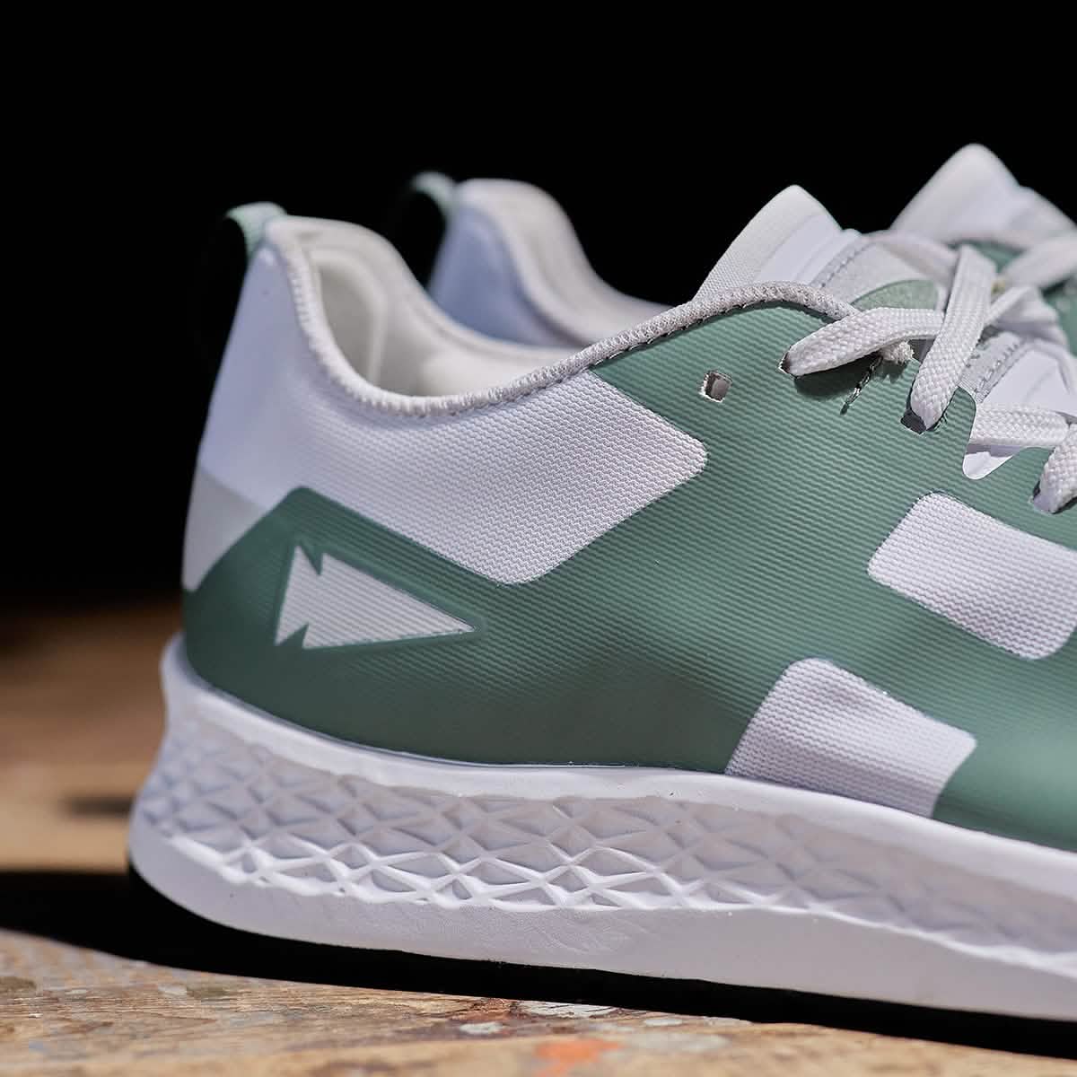 A close-up of the Women's Rough Runner - Seaspray sneaker by GORUCK highlights its green and white design. The shoe is adorned with a stylized lightning bolt symbol on the side and features a textured white sole equipped with a Gradient Density EVA Midsole. The dark background accentuates the intricate details of the shoe, making it stand out.