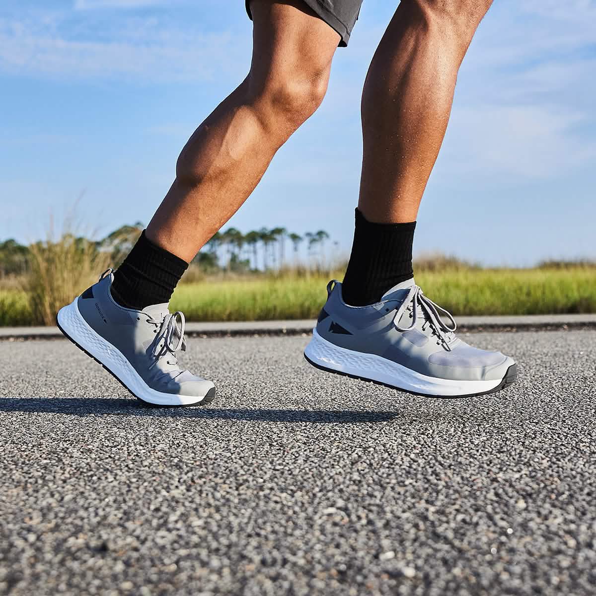 A person jogging on a paved road, channeling their inner Special Forces in GORUCK's Men's Rough Runner - Speed Grey + White shoes, black socks, and gray shorts. The backdrop showcases grassy fields and a clear blue sky, adding a scenic gradient density to the scene.