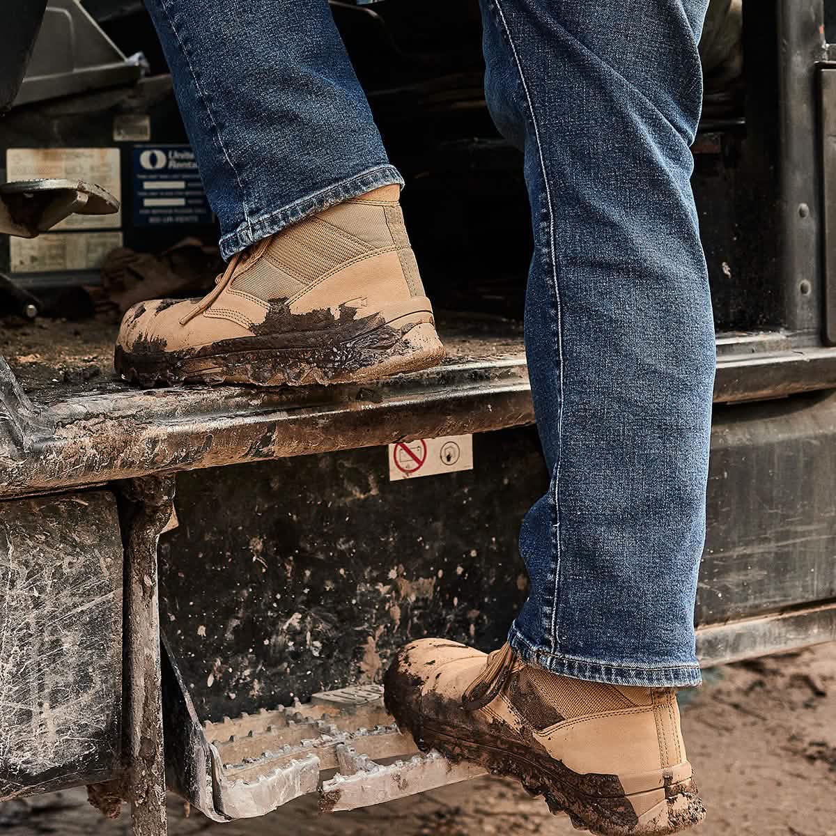 A person wearing the MACV-2 Safety Boot - Mid Top in tan by GORUCK and blue jeans steps onto a dirt-covered vehicle step. The background includes parts of the vehicle and a no-smoking sign.