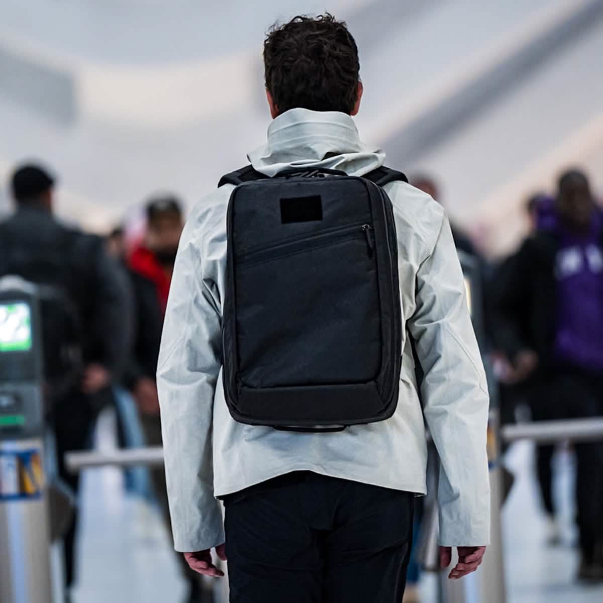 A person carrying a GR2 USA x Carryology - Ultra Black backpack, crafted from UHMWPE fabric, walks through a busy indoor area, seen from behind.