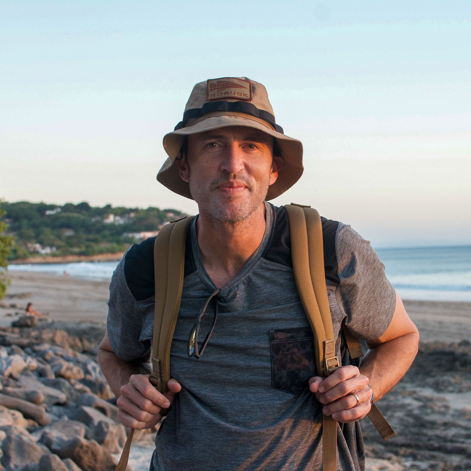 A man wearing a hat and carrying the Mesh Ruck stands on a rocky beach, with trees and the ocean in the background at sunset.