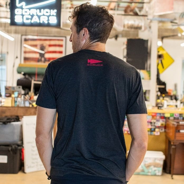 A man stands in an office, facing away, wearing a black Buddy FCKR Friday t-shirt beneath a neon GORUCK SCARS sign.